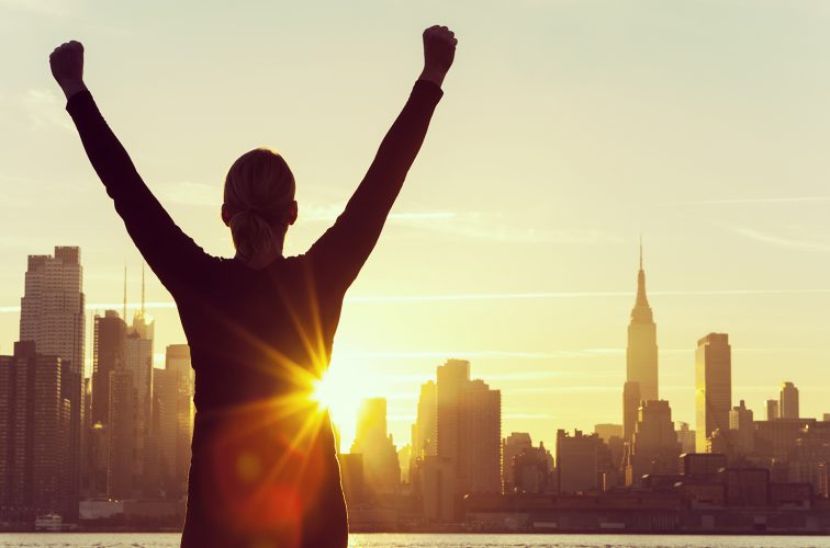 Silhouette of a successful woman or girl arms raised celebrating at sunrise or sunset in front of the New York City Skyline